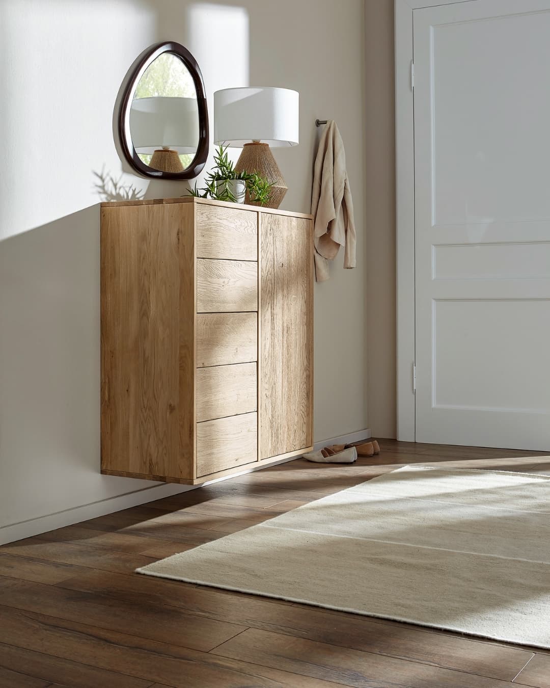 Moderne Holzkommode mit Spiegel, Lampe und Pflanzen in einem hellen Raum mit Teppich und offener Tür.