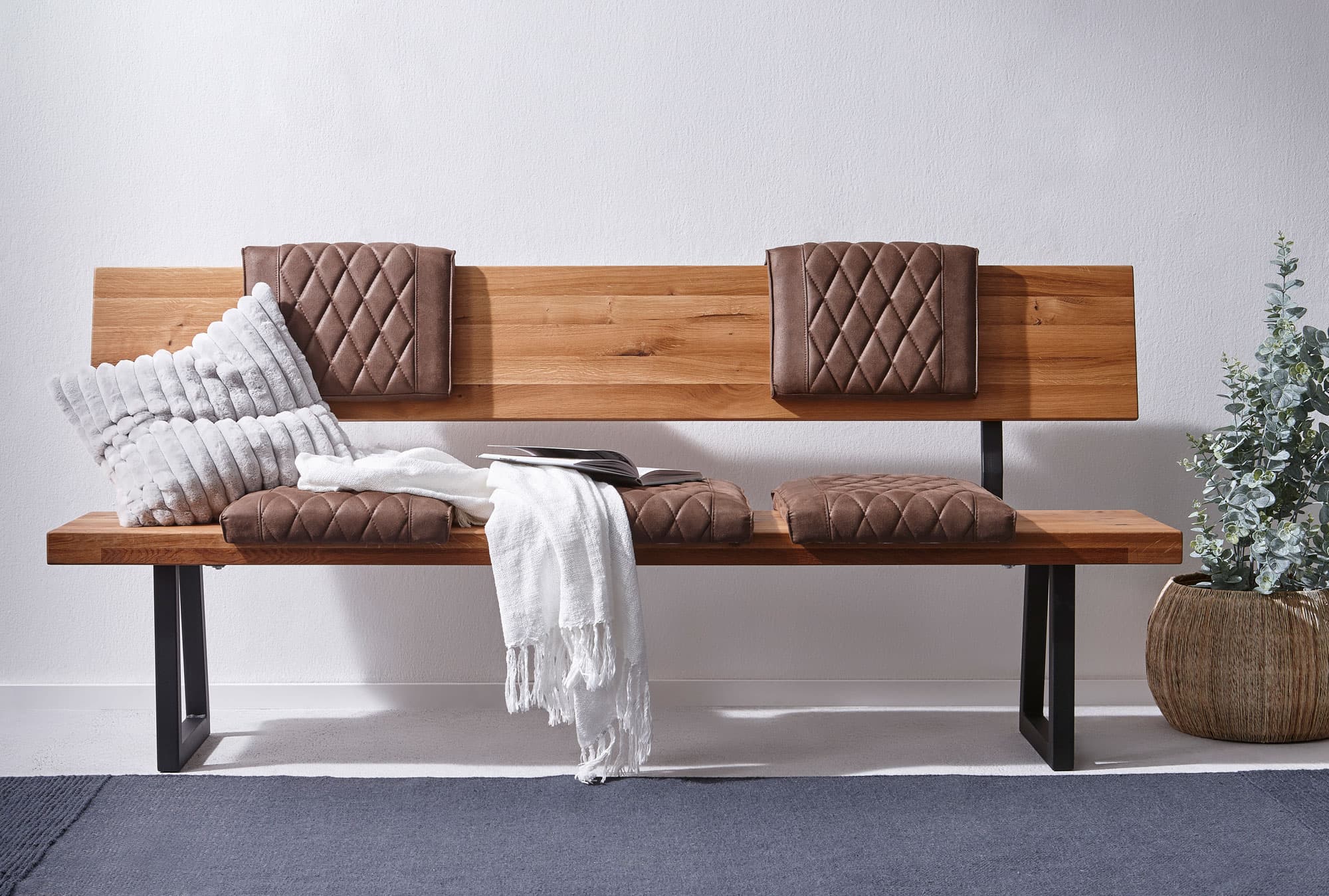 Holzbank mit Kissen, Decke und Pflanze in einem modernen Raum. Minimalistisches Design, warme Farben.