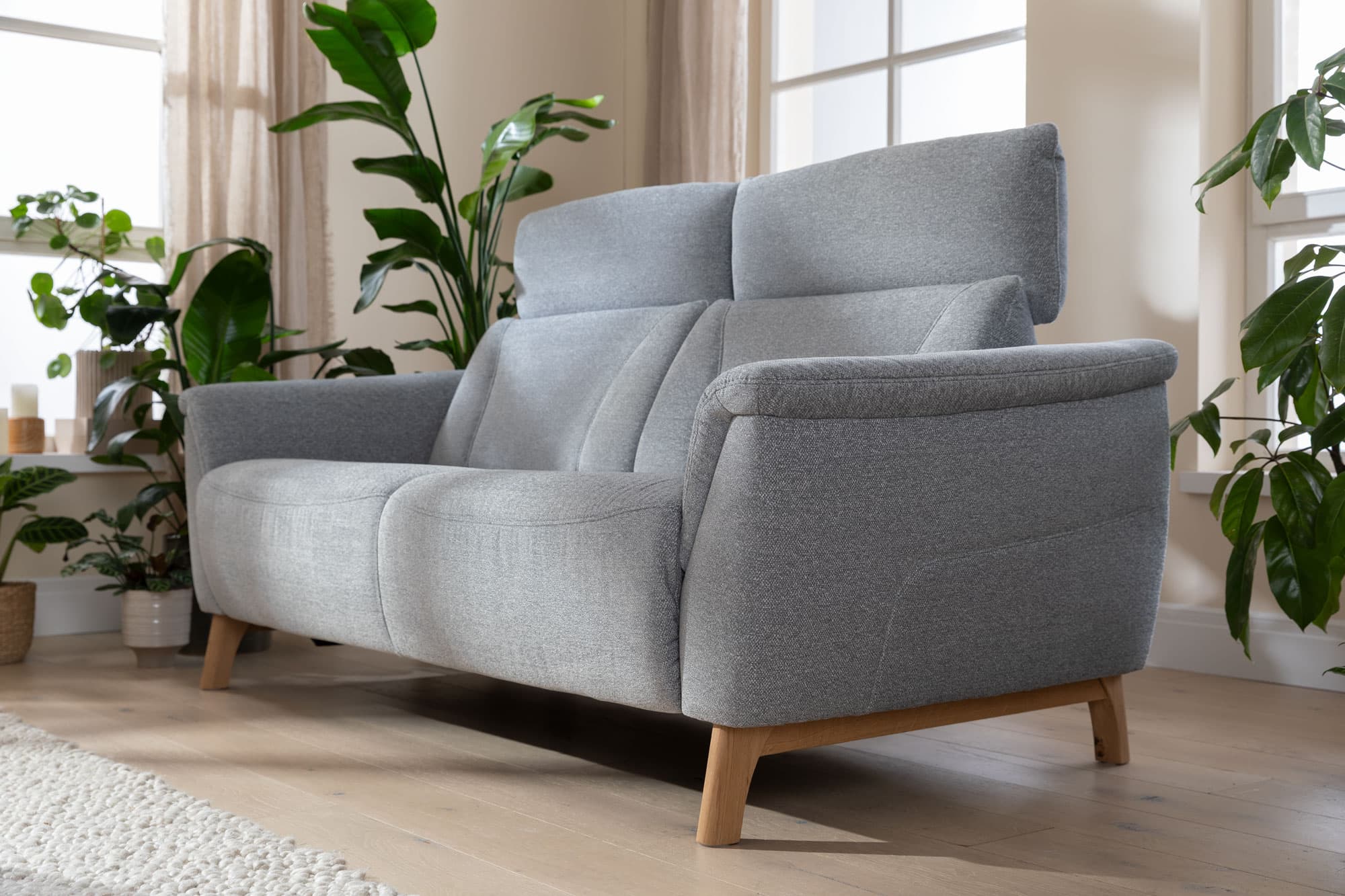 Gemütliches graues Sofa mit Holzfüßen, umgeben von Pflanzen und großen Fenstern in einem hellen Raum.