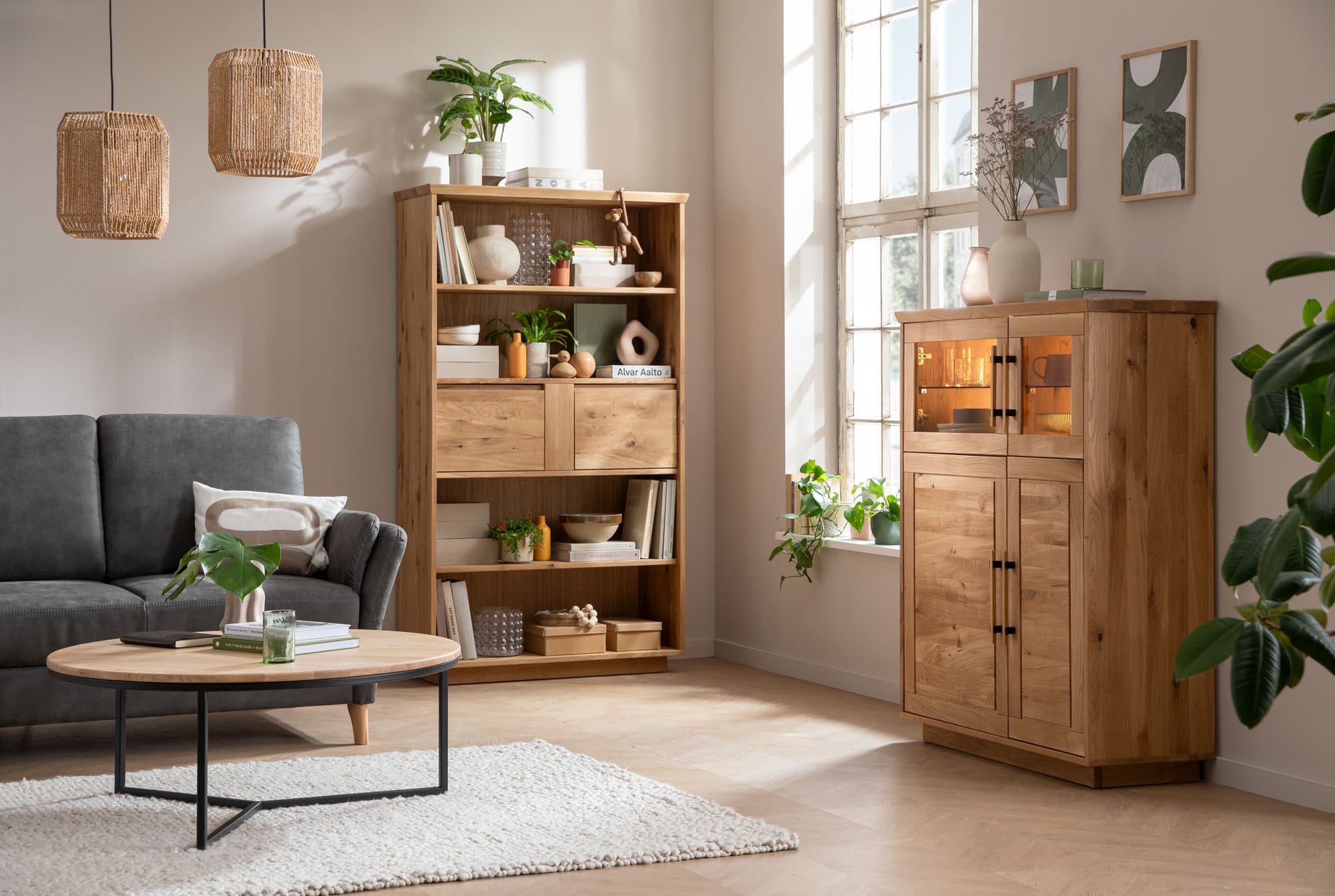 Ein gemütliches Wohnzimmer mit einem grauen Sofa, Holzmöbeln, einem runden Tisch und Pflanzen. Helle Fenster sorgen für Licht.