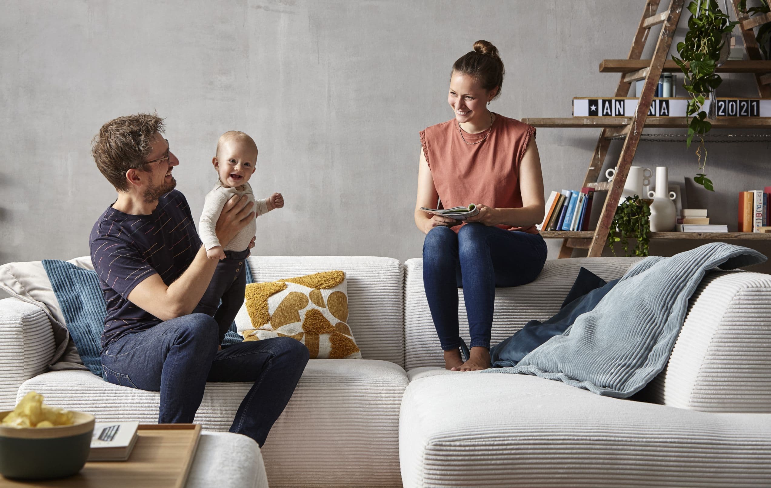 Ein Paar sitzt auf einem Sofa, der Mann hält ein Baby, während die Frau auf einem Tablet schaut. Umgebende Einrichtung ist modern.