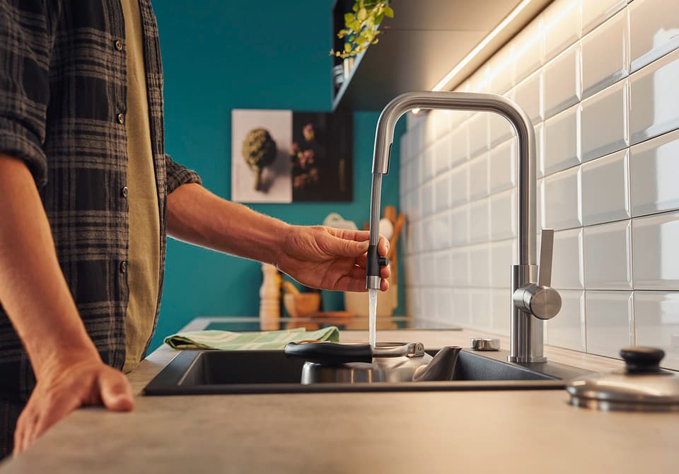 Eine Person steht vor einer modernen Küchenspüle und dreht den Wasserhahn auf. Im Hintergrund sind Küchendetails sichtbar.