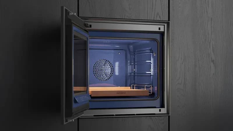Offene Mikrowelle mit grauem Inneren und gitterartigem Ventilator, eingebaut in eine schwarze Küchenwand.