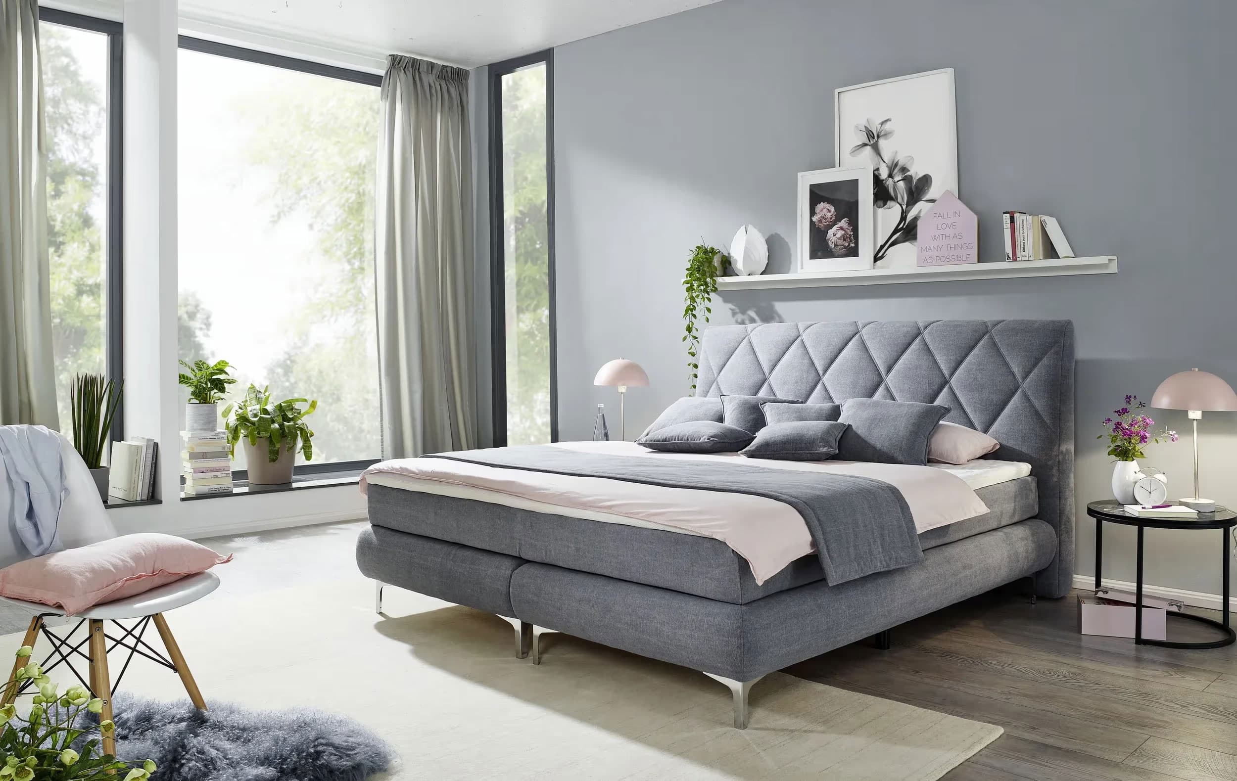 Modernes Schlafzimmer mit grauem Doppelbett, hellen Wänden, großen Fenstern und dekorativen Pflanzen.