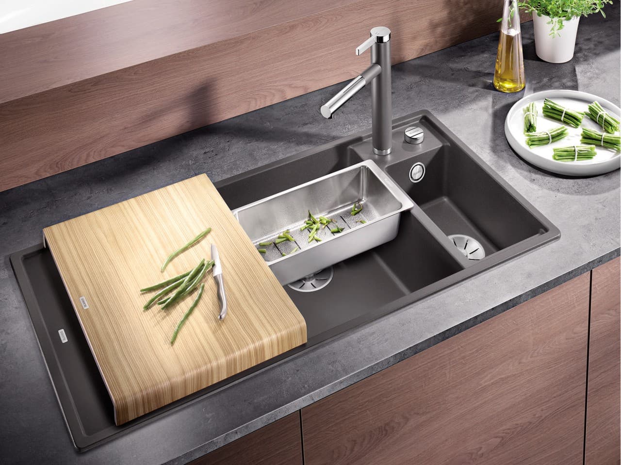 Moderne Spüle mit Holzschneidebrett, Edelstahl-Geschirr und frischen Kräutern auf einer Arbeitsplatte aus dunklem Material.