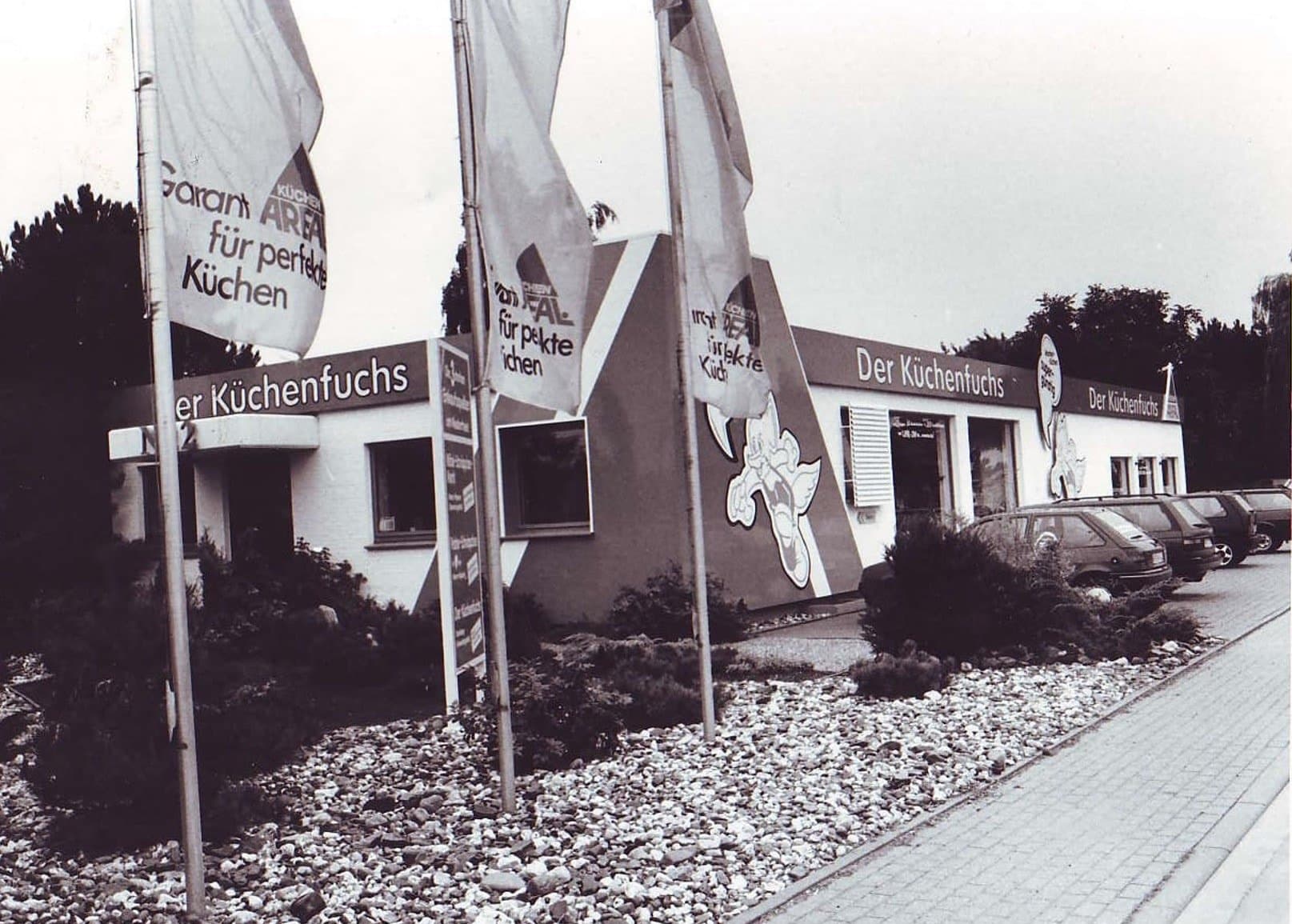 Fassade eines Geschäfts mit mehreren Fahnen, Gartenlandschaft und Schotterweg im Vordergrund.