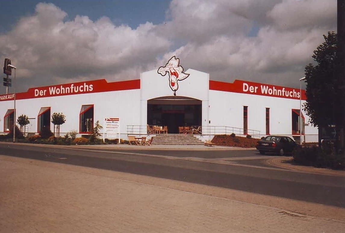 Eine große, weiße Verkaufsstätte mit rotem Schriftzug "Der Wohnfuchs" und umgebenden Bäumen unter cloudy sky.
