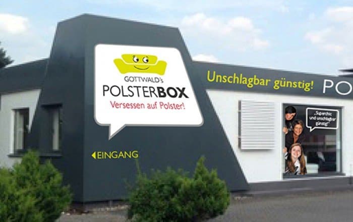 Moderne Gebäude mit Schild "POLSTERBOX" und Eingangsbereich, umgeben von grünen Pflanzen.