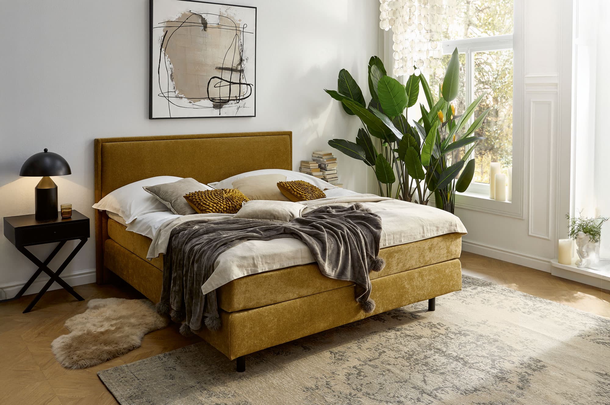 Gemütliches Schlafzimmer mit einem senfgelben Bett, Kissen, einer Decke, Pflanzen und einem modernen Kunstwerk.