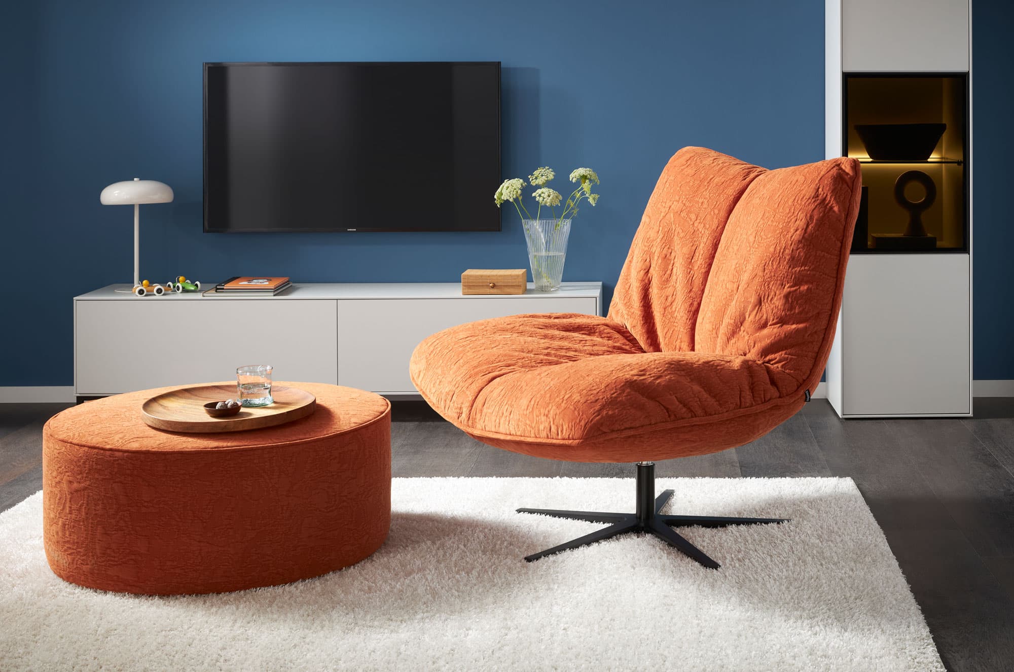 Oranger Sessel mit niedrigem Tisch auf einem Teppich vor blauer Wand und TV-Möbel. Minimalistische, moderne Einrichtung.