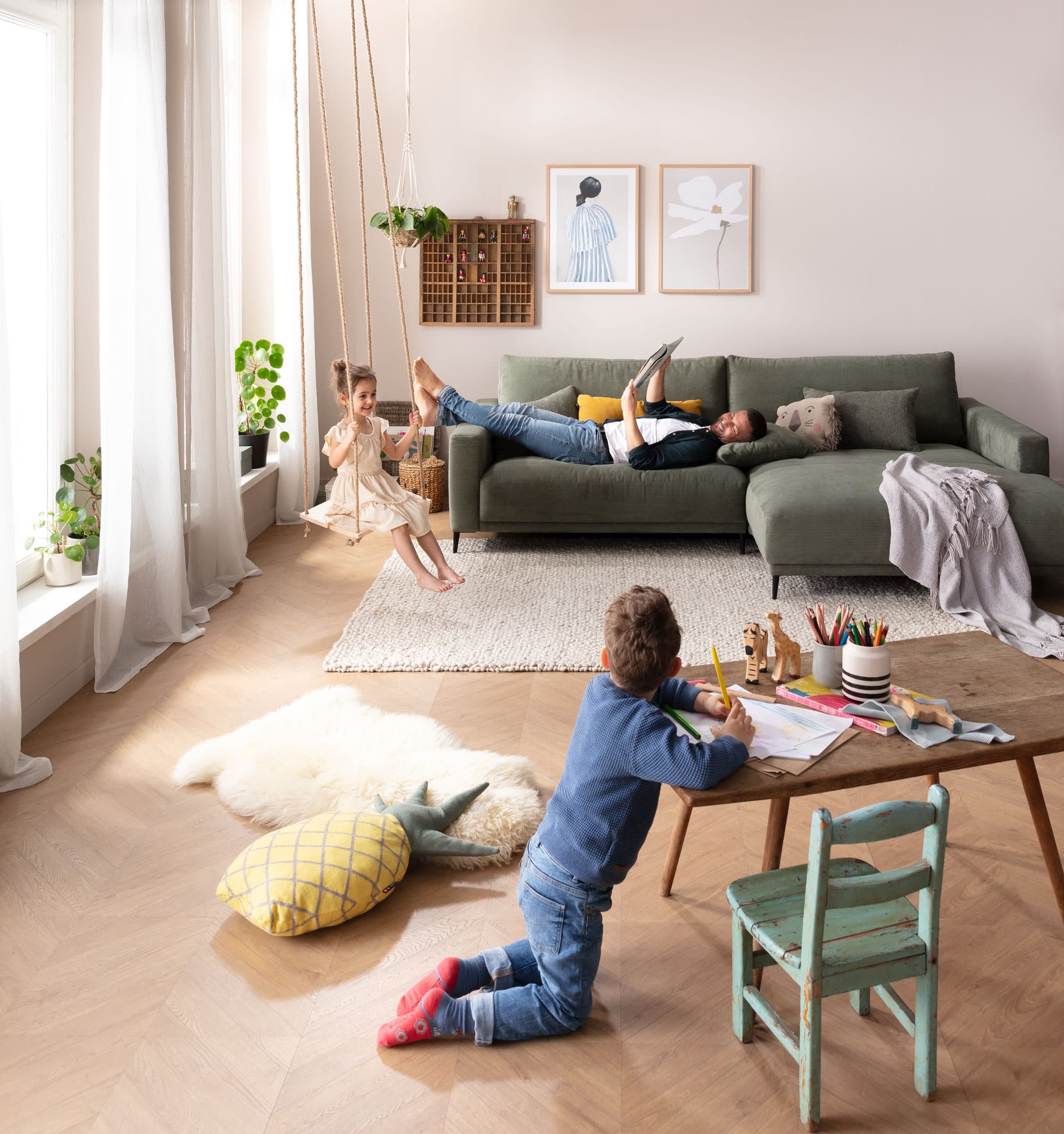 Helles Wohnzimmer mit Kind, das am Tisch malt, während eine Frau auf dem Sofa entspannt und ein anderes Kind schaukelt.