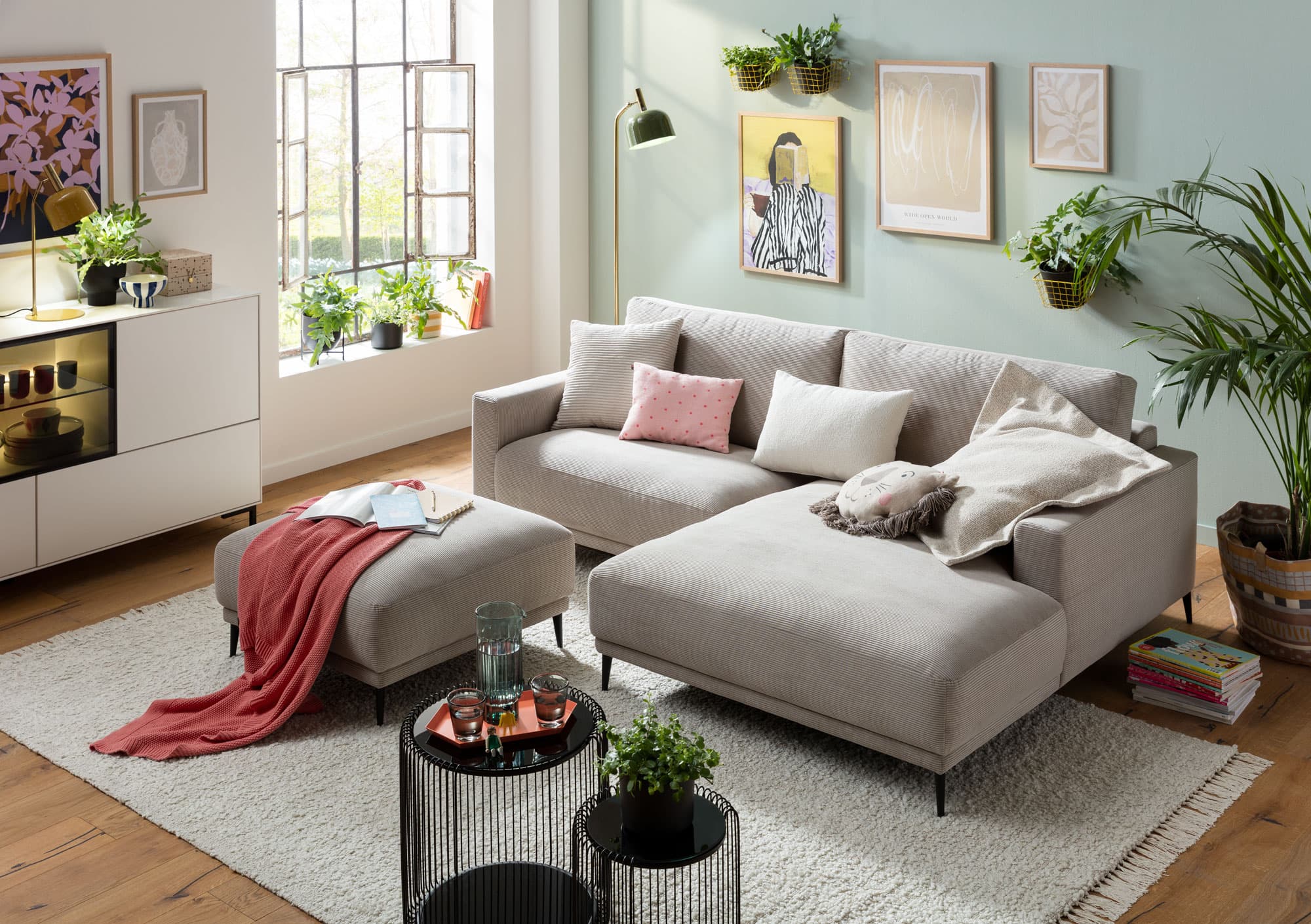 Helles Wohnzimmer mit grauem Sofa, bunten Kissen, Pflanzen und modernen Wandbildern. Holzfußboden und stilvolle Dekoration.