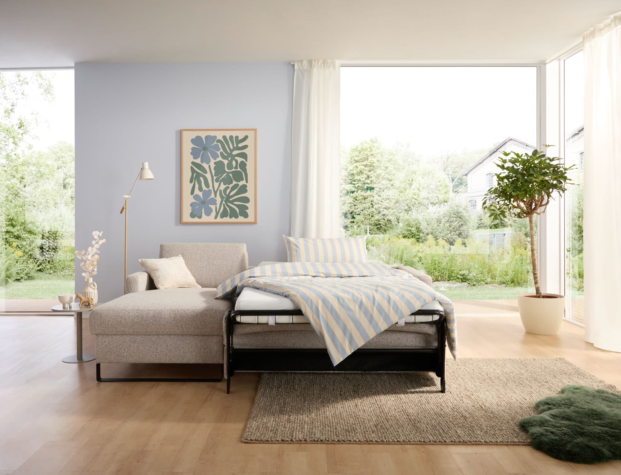 Helles Zimmer mit einem Schlafsofa, einer Fensterfront, einem Kunstwerk an der Wand und Pflanzen im Hintergrund.