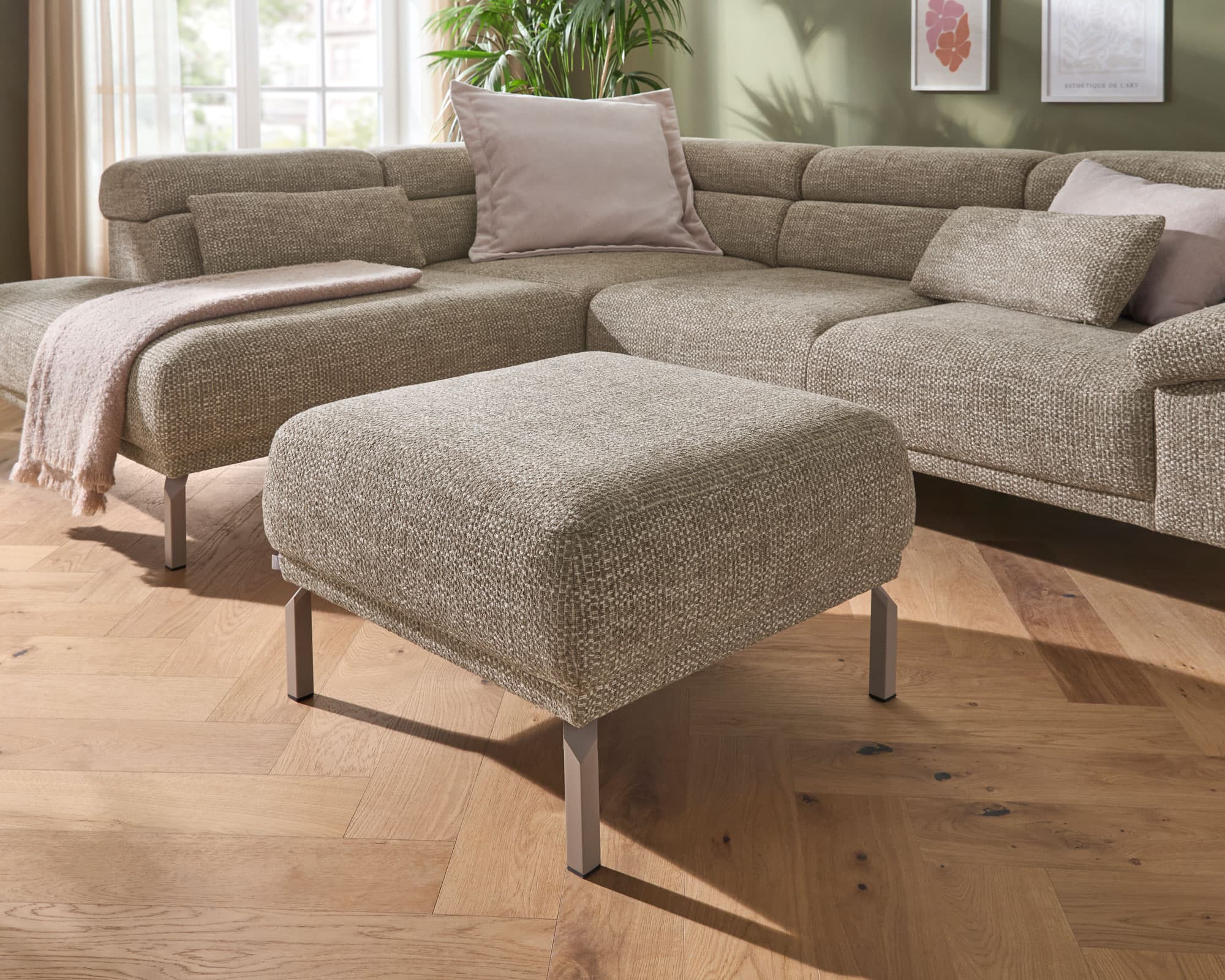 Beige Couch mit Kissen und Hocker auf Holzboden, umgeben von großen Fenstern und einer grünen Wand.