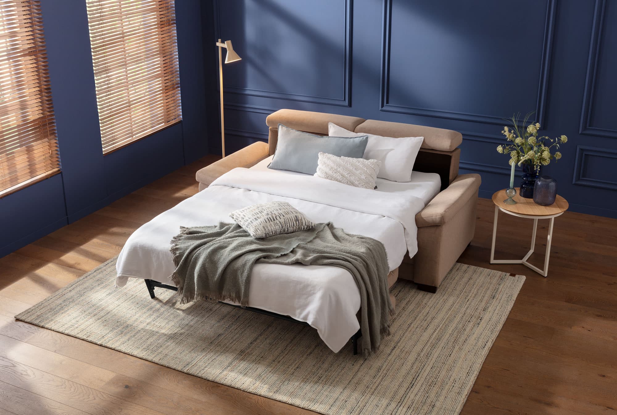 Elegantes Schlafzimmer mit einem ausziehbaren Sofa, Kissen, einer Decke und einem Beistelltisch mit Blumen. Holzfußboden und Fenster.
