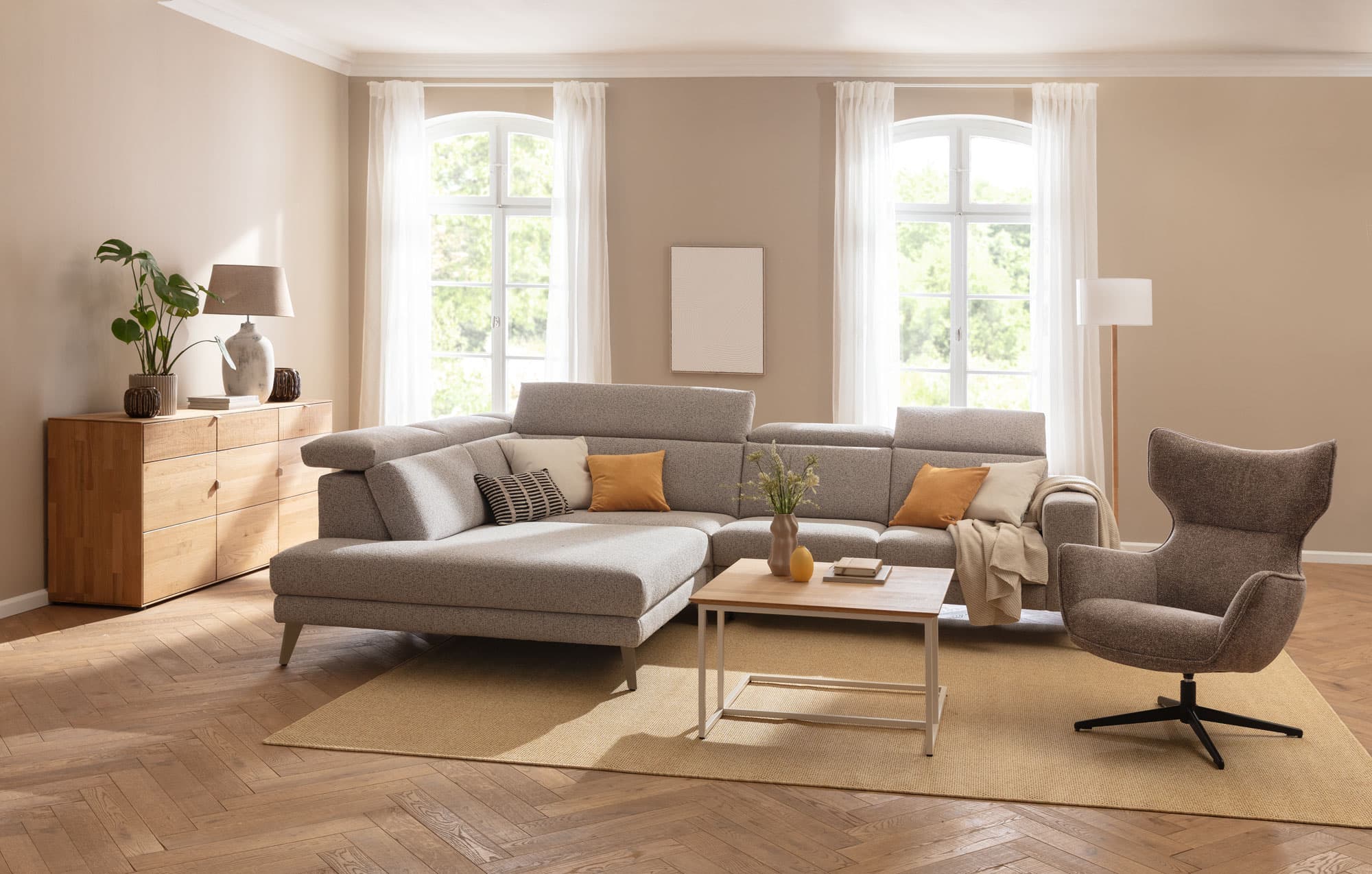 Helles Wohnzimmer mit grauem Sofa, Kaffeetisch, Sessel, Holzkasten und Pflanzen vor großen Fenstern.